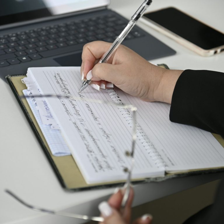 Main mettant à jour des documents sur un carnet, avec des lunettes et un téléphone à côté.