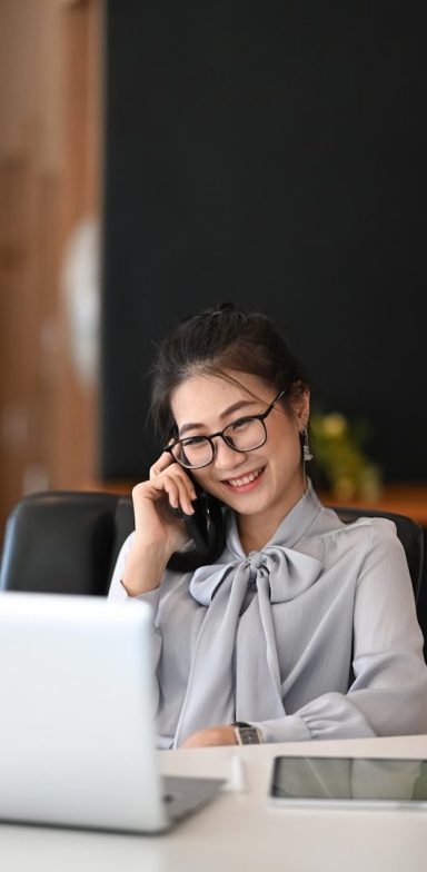 Femme souriante en chemisier, parlant au téléphone, assise devant un ordinateur portable.