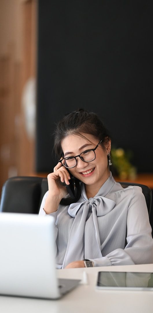 Femme souriante en chemisier, parlant au téléphone, assise devant un ordinateur portable.