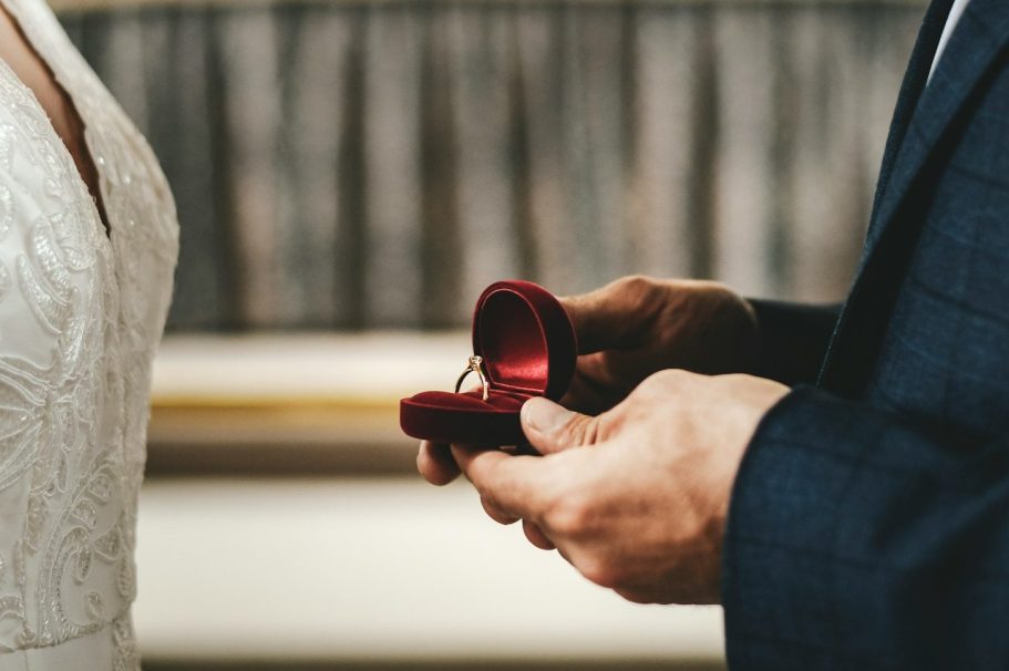 Main dans une boîte rouge, tenant une bague, avec une mariée en arrière-plan.