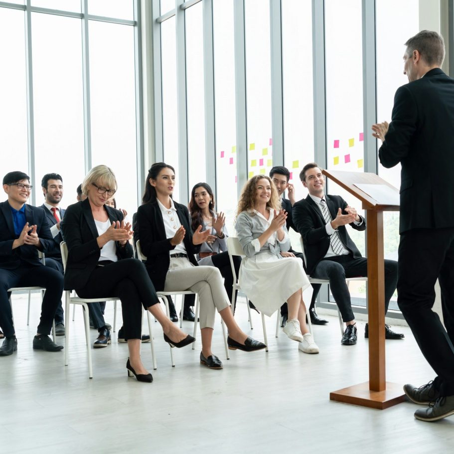 Un orateur présente devant un public applaudissant dans une salle lumineuse.
